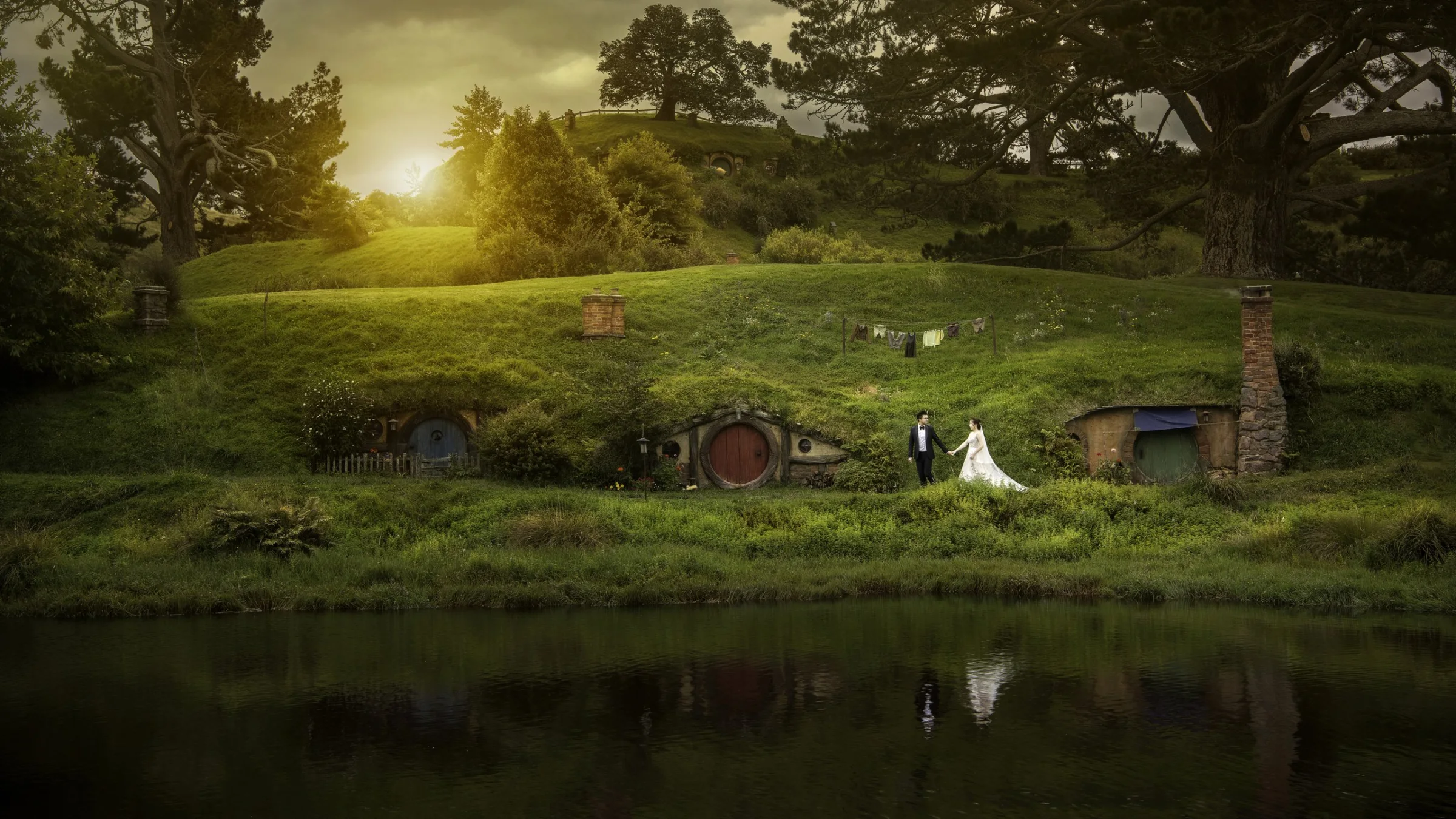 Bride and groom walking hand in hand past the round-door hillside homes at Hobbiton, New Zealand — pre-wedding by Kenn Photographic Arts