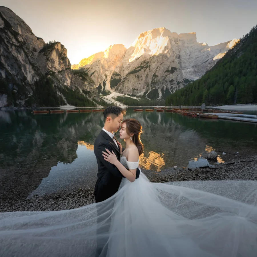 Mable & Hon embracing on the shore of Lago di Braies at sunrise, her long veil across the pebbles — Dolomites pre-wedding by Kenn Photographic Arts