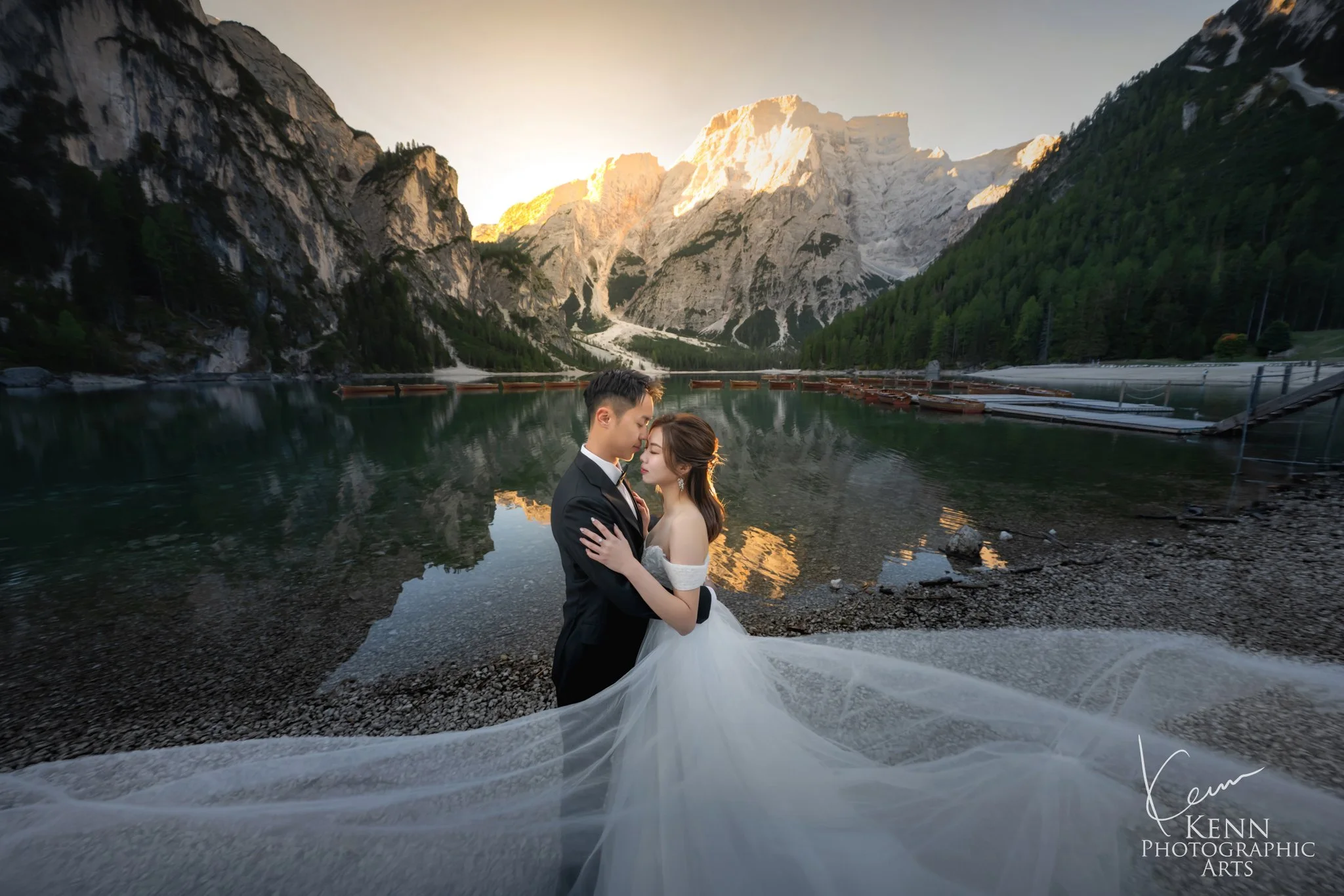 Mable & Hon embracing on the shore of Lago di Braies at sunrise, her long veil across the pebbles — Dolomites pre-wedding by Kenn Photographic Arts
