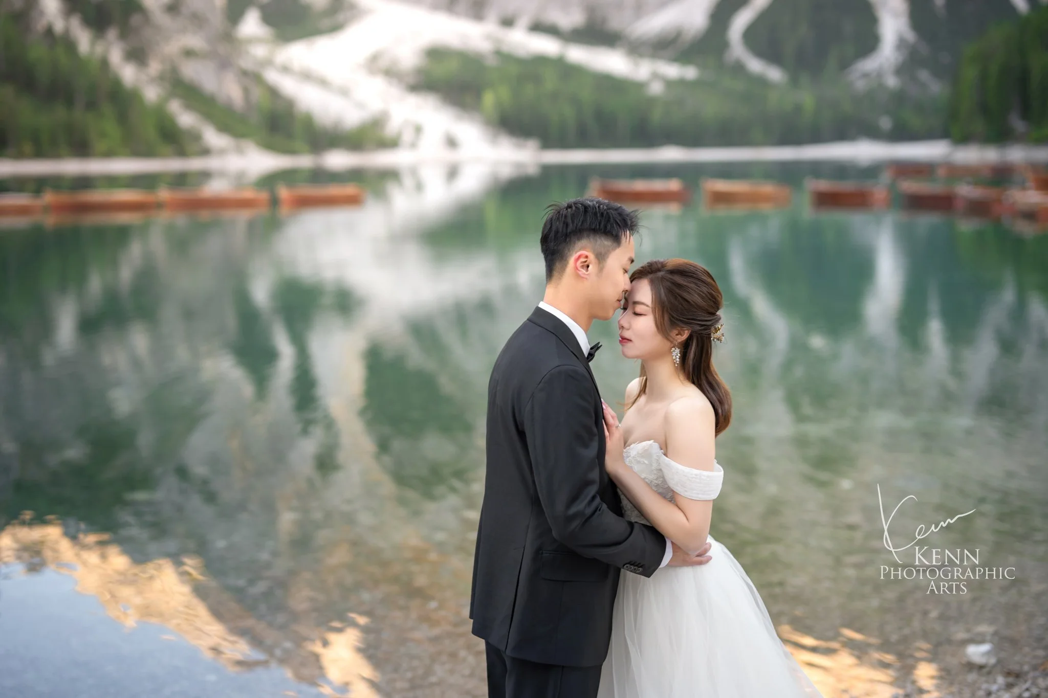Mable & Hon in a close embrace with the green lake reflection behind them — Dolomites pre-wedding by Kenn Photographic Arts