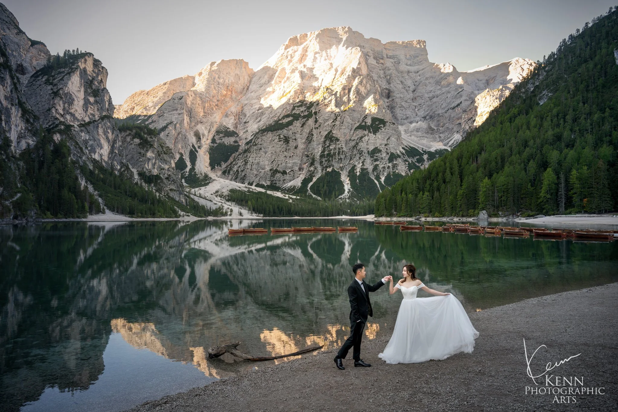 Groom leading the bride along the shore of Lago di Braies, mountains mirrored in the lake — Dolomites pre-wedding by Kenn Photographic Arts