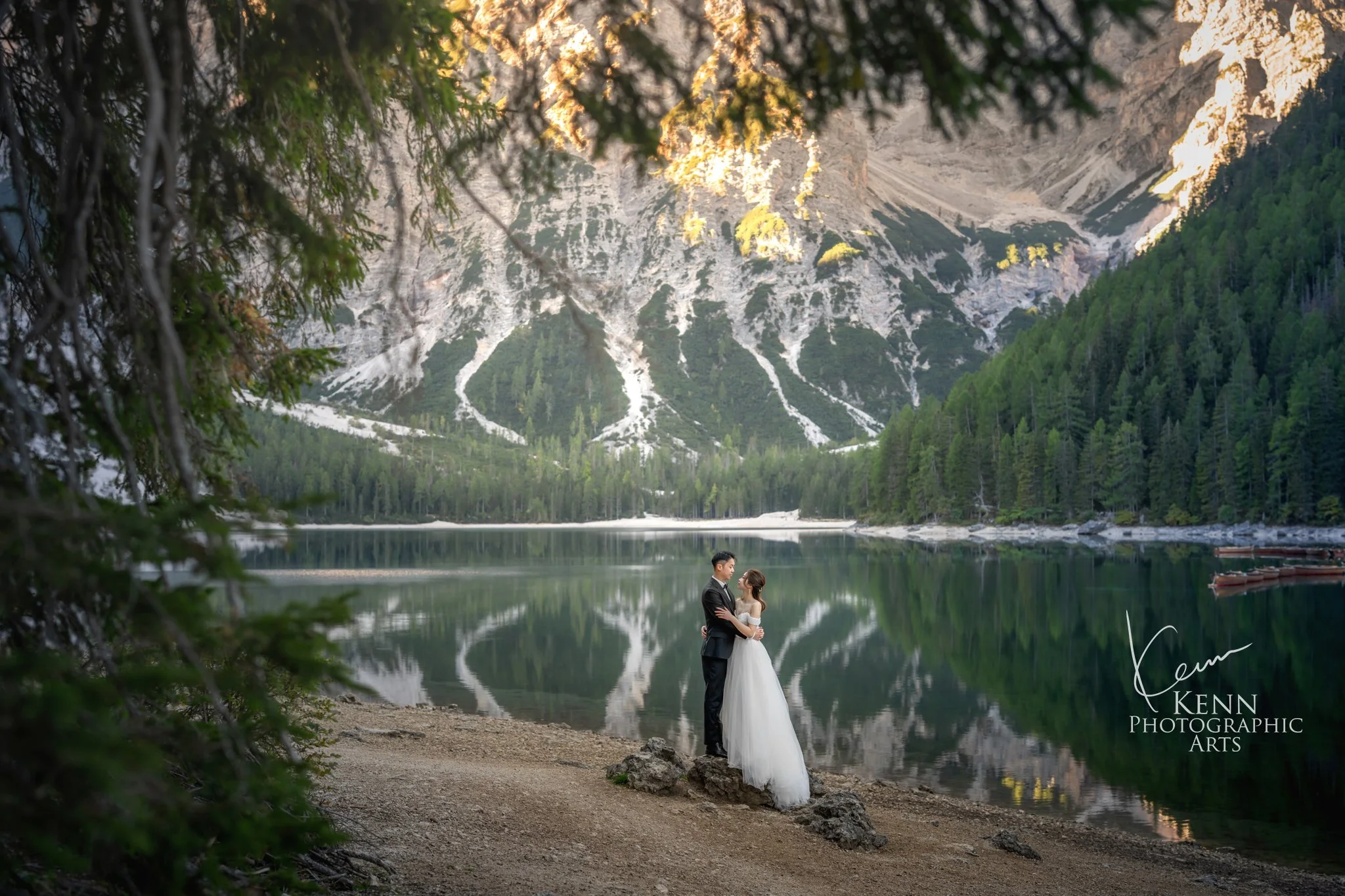 Mable & Hon framed by pine branches at the edge of the lake — Dolomites pre-wedding by Kenn Photographic Arts