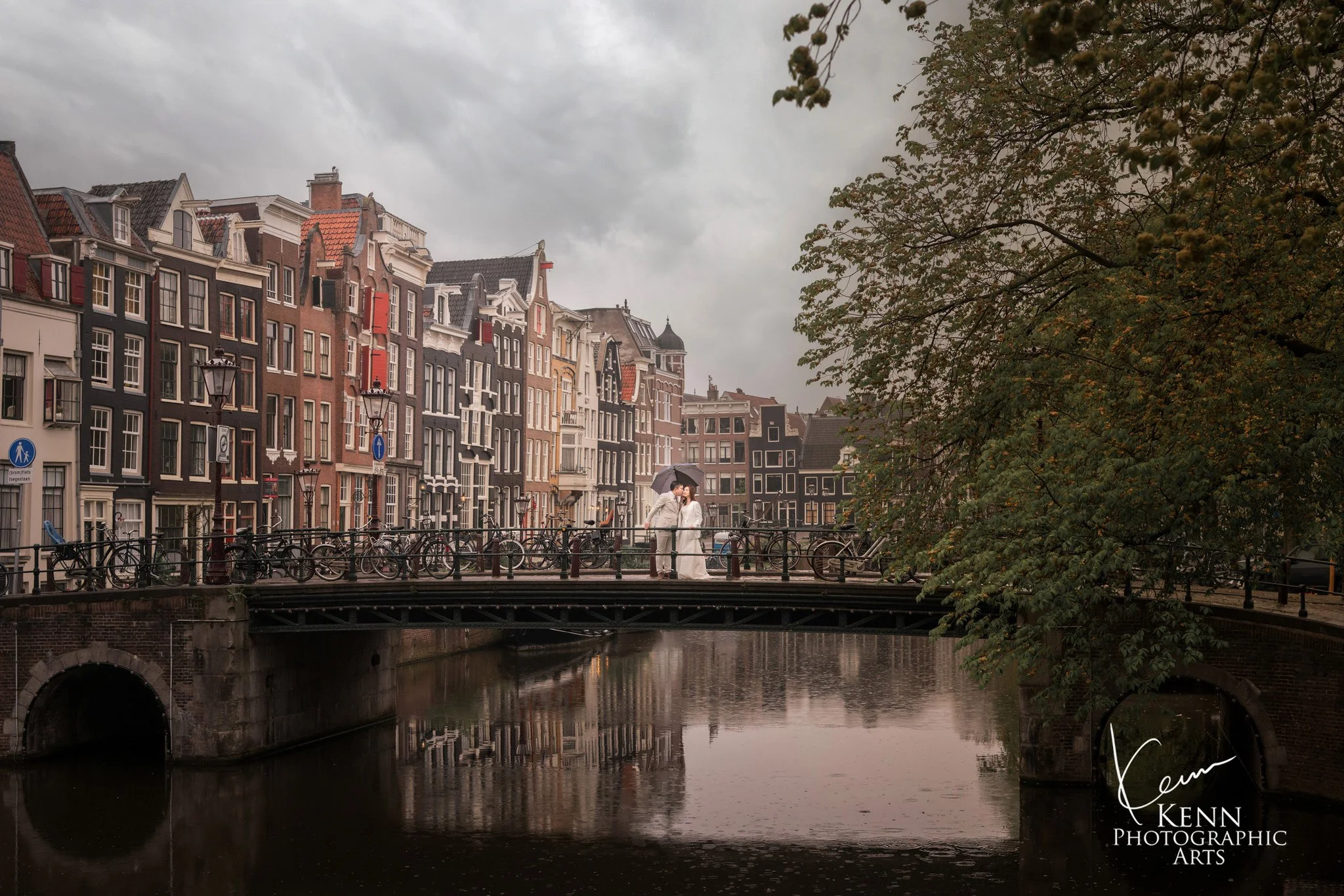 Michelle & Vincent on a canal bridge at dawn, Amsterdam's gabled houses reflected in the water — Netherlands pre-wedding by Kenn Photographic Arts