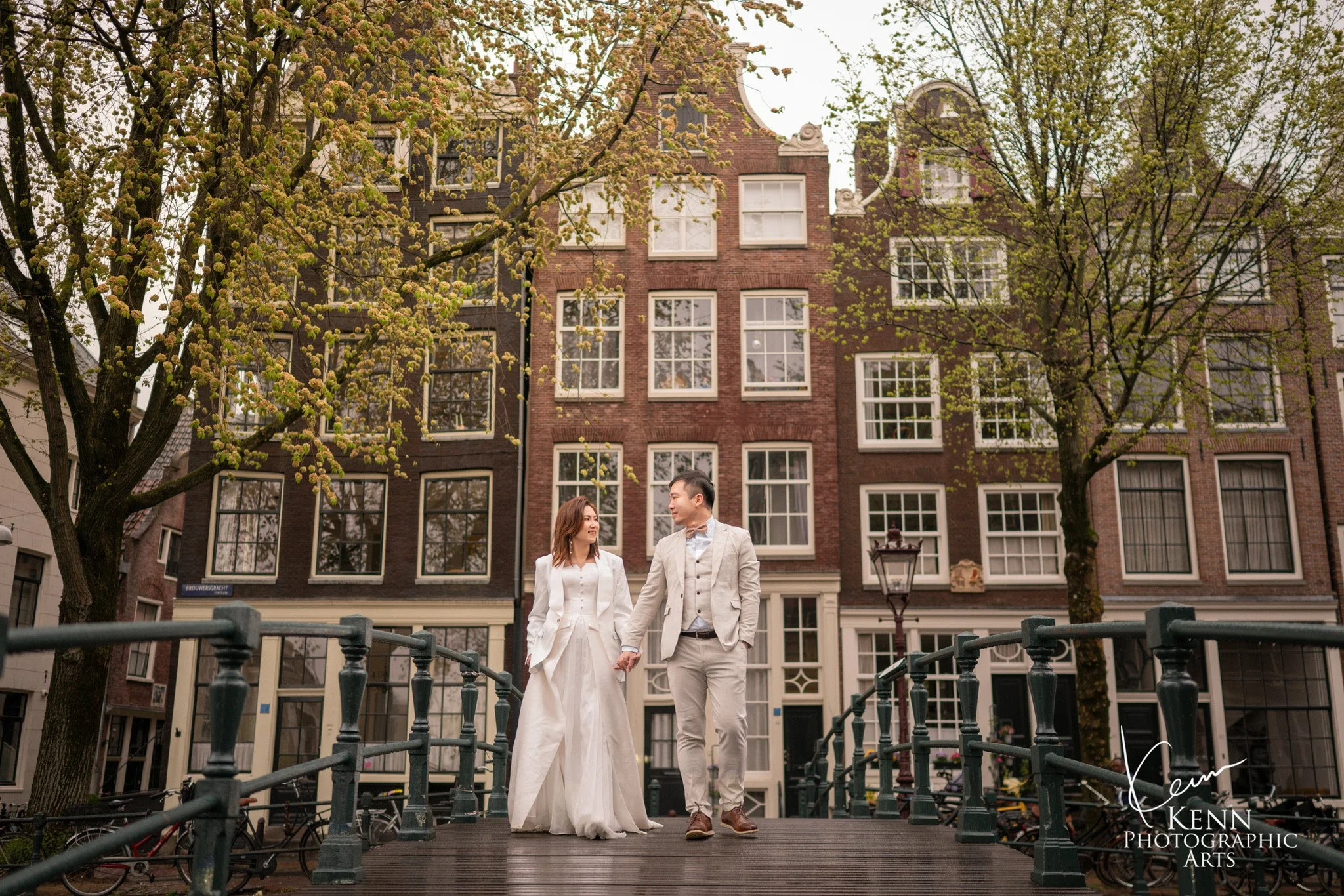 Michelle & Vincent walking across a canal bridge in front of Amsterdam's brick canal houses — Netherlands pre-wedding by Kenn Photographic Arts