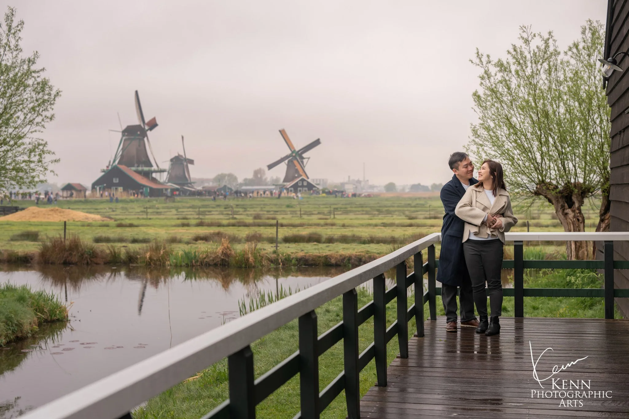 Michelle & Vincent on a wooden boardwalk with the windmills of Zaanse Schans behind — Netherlands pre-wedding by Kenn Photographic Arts