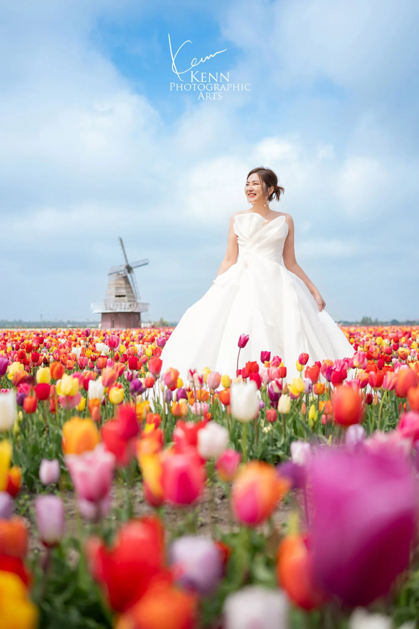Bride standing in a tulip field with a windmill on the horizon — Netherlands pre-wedding by Kenn Photographic Arts